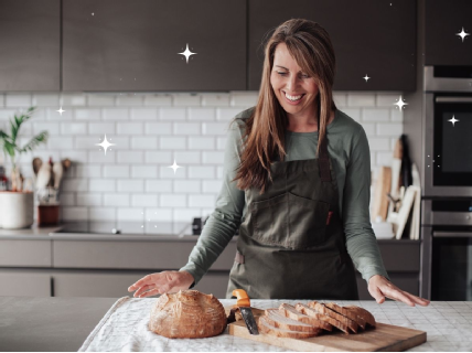 Author Laura Strange standing in her kitchen, looking at a freshly made loaf of gluten free bread placed on a wooden bread board.